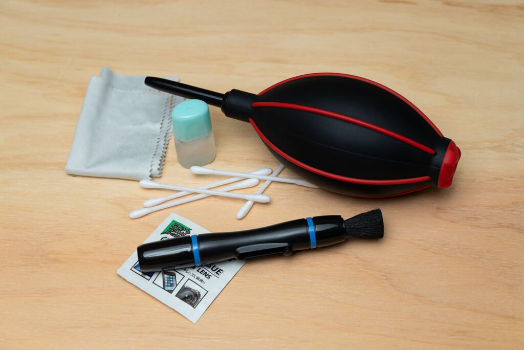 Camera lens cleaning tools including air blower, lens pen, microfiber cloth and cleaning solution on wooden surface